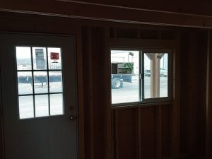 Inside the Lofted Dutch Cabin - 1456, an unfinished wooden interior includes a door and a window. Outside, a trailer and vehicle can be seen on the paved area through the window. The limited light entering creates a striking contrast between the dim lofted interior and the bright exterior.