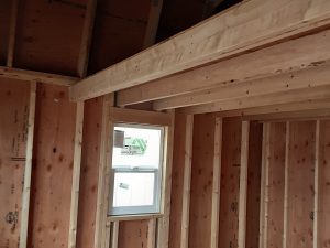 The interior of the Lofted Dutch Cabin - 1456 under construction showcases its exposed wooden beams and framing. A small window adorns the wall, while unfinished plywood covers both the walls and ceiling, emphasizing the cabin's rustic charm.