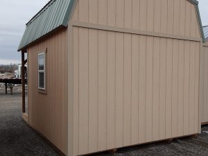 A compact Lofted Dutch Cabin - 1456, featuring a tan exterior and a green metal roof, with one window situated on the left side. Positioned on a gravel surface outdoors, it is surrounded by other partially visible structures in the background.