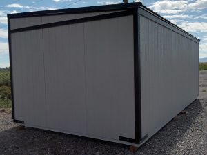 A large, rectangular gray Buckaroo - 1951 storage container with black trim sits on a gravel ground under a clear sky. In the background, an electric pole with wires adds a touch of nostalgia to this scene reminiscent of the sturdy structures from that era.