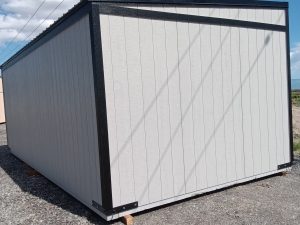 Under a partly cloudy sky, the Buckaroo - 1951 stands on a gravel surface, embodying rugged simplicity. Its large rectangular shape features plain light gray walls with black trim, typical of its style, while power lines stretch across the background. The shed has no visible windows or doors.