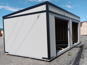 A partially constructed Buckaroo - 1951 white and gray rectangular shed is situated on a gravel lot under a blue sky with occasional clouds. The framed openings hint at future windows or doors. Additional structures in the background enhance the rustic charm of the scene.