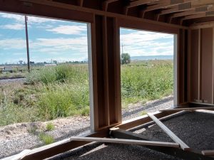 Looking through two expansive window frames inside the unfinished building, a scene unfolds of a grassy field and power lines beneath a partly cloudy sky, evoking the nostalgia of Buckaroo - 1951. The natural light bathes the wooden framework and gravel floor, infusing this modern space with rustic charm.
