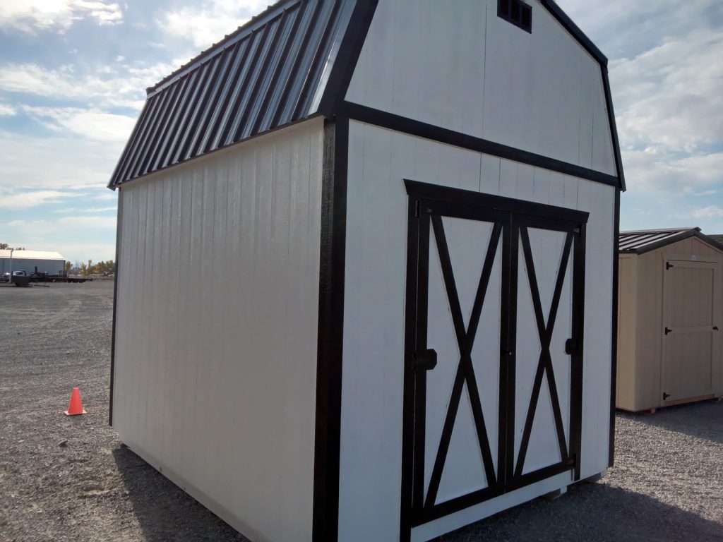 The Lofted Dutch Barn - 2114, featuring a white exterior with a black metal roof and trim and double barn-style doors, is situated on a gravel lot beside a small orange traffic cone. In the background, another smaller shed is visible.