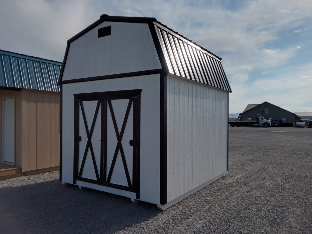 The **Lofted Dutch Barn - 2114**, a compact white storage shed with a black barn-style roof and lofted design, features double doors and stands on a gravel surface. The partly cloudy morning sky frames the structure, with another building visible in the background.