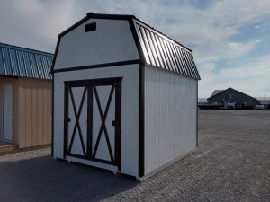 The **Lofted Dutch Barn - 2114**, a compact white storage shed with a black barn-style roof and lofted design, features double doors and stands on a gravel surface. The partly cloudy morning sky frames the structure, with another building visible in the background.