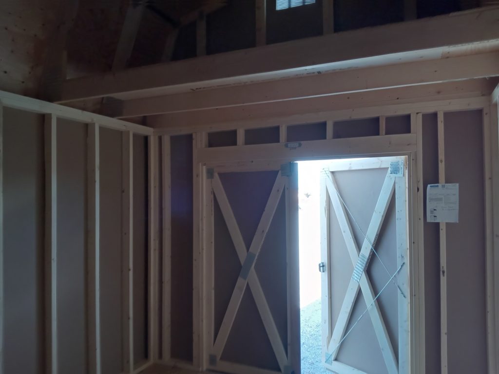 Interior view of a Lofted Dutch Barn - 2114 wooden shed under construction. The door is partially open, allowing light to fill the dim space. Exposed wooden frames and walls are visible, with a note attached to the open door.