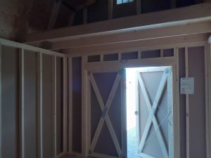 Interior view of a Lofted Dutch Barn - 2114 wooden shed under construction. The door is partially open, allowing light to fill the dim space. Exposed wooden frames and walls are visible, with a note attached to the open door.