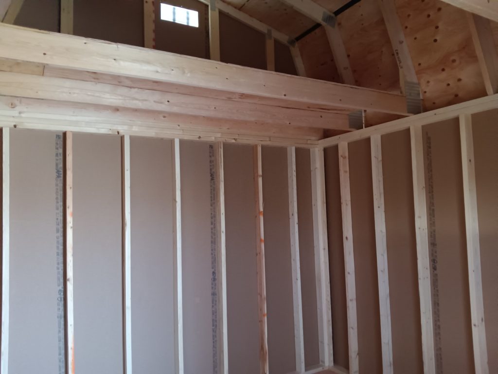 Interior view of the Lofted Dutch Barn - 2114 under construction. The image features exposed wooden beams and studs, along with partially installed drywall. A small window near the top allows natural light to illuminate this rustic space, showcasing the craftsmanship involved in creating this barn.