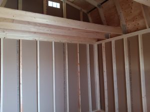 Interior view of the Lofted Dutch Barn - 2114 under construction. The image features exposed wooden beams and studs, along with partially installed drywall. A small window near the top allows natural light to illuminate this rustic space, showcasing the craftsmanship involved in creating this barn.