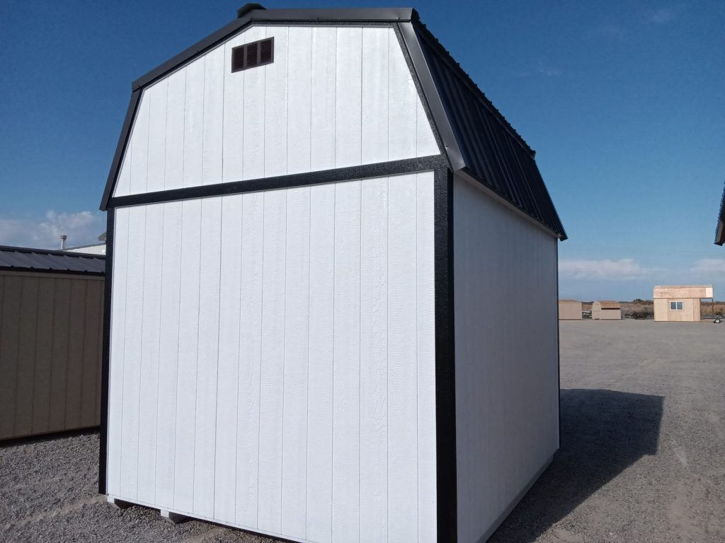 A Lofted Dutch Barn - 2114, featuring a white structure with a tall profile and accented by a black roof and trim, stands on a gravel lot. It showcases a classic design with a large door at the front. In the background, similar barns can be seen under a clear blue sky.