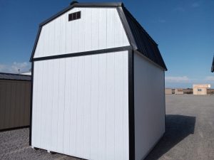 A Lofted Dutch Barn - 2114, featuring a white structure with a tall profile and accented by a black roof and trim, stands on a gravel lot. It showcases a classic design with a large door at the front. In the background, similar barns can be seen under a clear blue sky.