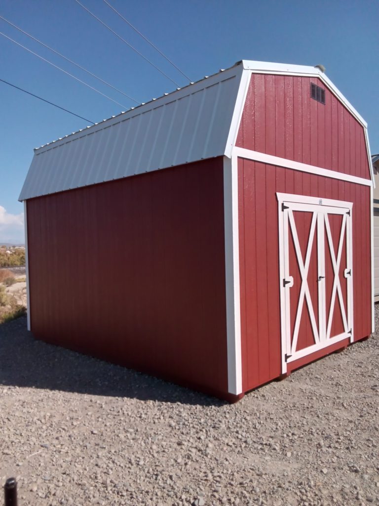 A Lofted Dutch Barn - 2113 shed featuring a white roof and trim rests on a gravel surface with its doors sealed, all beneath a clear blue sky. Power lines trace the horizon in the background, enhancing the rural charm.