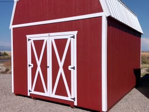 A Lofted Dutch Barn - 2113, featuring white doors and trim, rests on a gravel surface beneath the clear blue sky. Its charming peaked roof stands out as power lines trace the background.