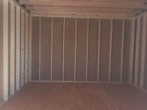The image features the interior of a Lofted Dutch Barn - 2113 currently under construction. The walls are framed with exposed beams, while sunlight streams through an open door, creating charming shadows on the plywood floor.
