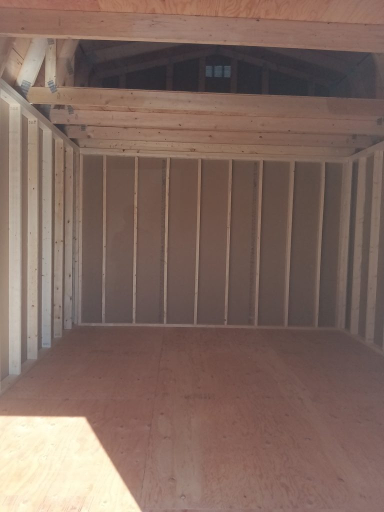 The image features the interior of a Lofted Dutch Barn - 2113 currently under construction. The walls are framed with exposed beams, while sunlight streams through an open door, creating charming shadows on the plywood floor.