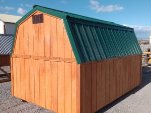 The Mini-Barn - 2306, featuring a wooden structure and a green, sloped metal roof, is situated on a gravel surface beneath a clear blue sky.