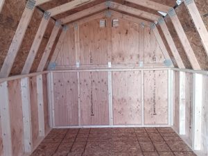 Inside the Mini-Barn - 2306, a peaked roof is supported by exposed beams, while unfinished wooden walls and floors exude rustic charm. The bright lighting accentuates the natural grain and texture of the wood, evoking a sense of simpler times in its serene emptiness.
