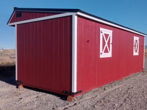 A red, barn-style shed with white trim and cross-braced windows sits on a gravel surface under a clear blue sky, reminiscent of the idyllic scene captured by the product Horseman - 2303.