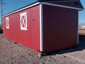 A Horseman - 2303 storage shed, featuring a red structure with white trim and two X-pattern windows reminiscent of a horseman's stable, stands on a gravel surface. The clear blue sky is set against power lines weaving through the backdrop.