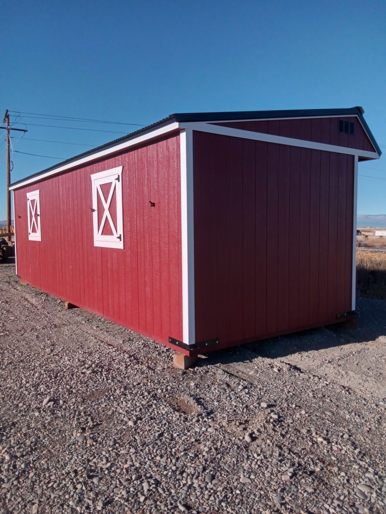 A Horseman - 2303 storage shed, featuring a red structure with white trim and two X-pattern windows reminiscent of a horseman's stable, stands on a gravel surface. The clear blue sky is set against power lines weaving through the backdrop.
