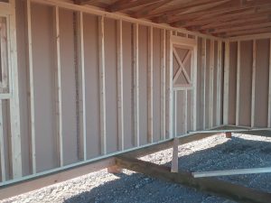 The partially constructed wooden structure, known as the Horseman - 2303, resembles a rustic horseman's shelter with its exposed framing and beams set on a gravel foundation. Its unfinished walls and visible wooden rafters allow natural light to stream through small window openings.