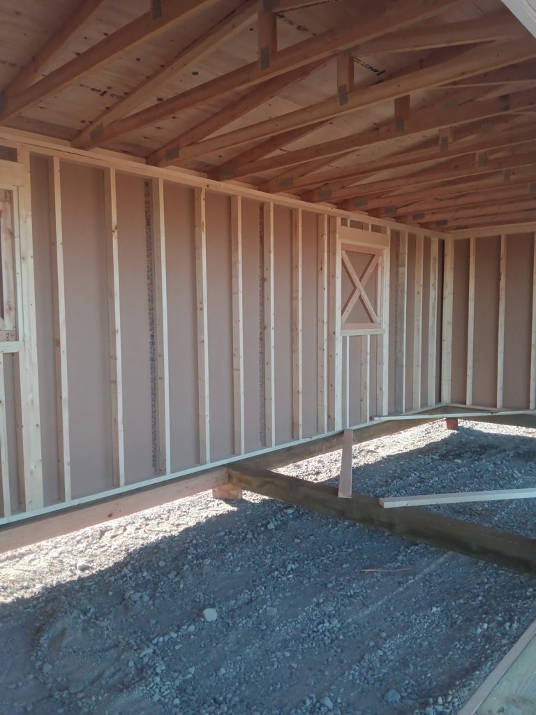 The partially constructed wooden structure, known as the Horseman - 2303, resembles a rustic horseman's shelter with its exposed framing and beams set on a gravel foundation. Its unfinished walls and visible wooden rafters allow natural light to stream through small window openings.