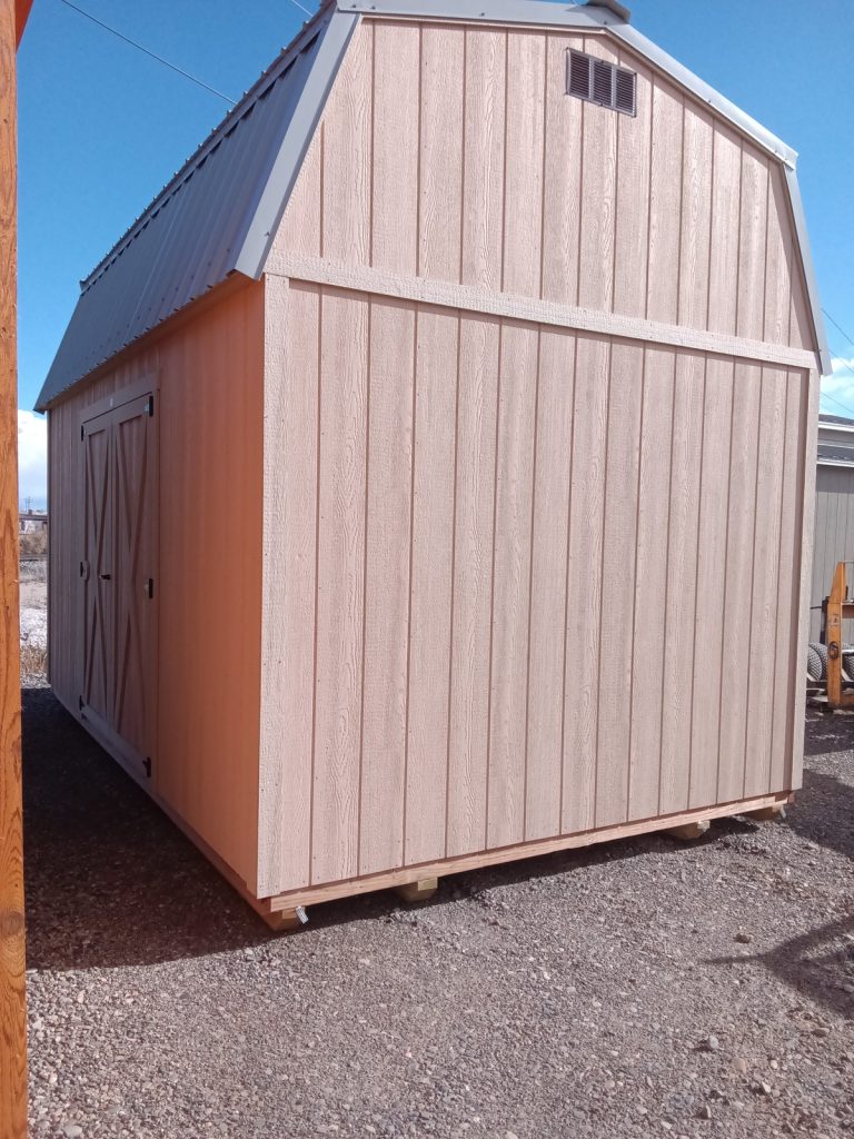 The Lofted Dutch Barn - 2368, featuring a crossbeam door and metal roof, is a charming structure in beige wood. It stands elegantly on a gravel surface beneath the clear blue sky.