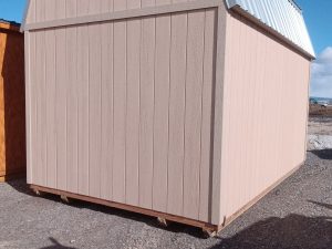 A Lofted Dutch Barn - 2368 featuring a tan wooden facade and a silver metal roof sits on a gravel surface beneath a clear blue sky. Shadows of a fence elegantly stretch across the ground in front, enhancing its rustic charm.