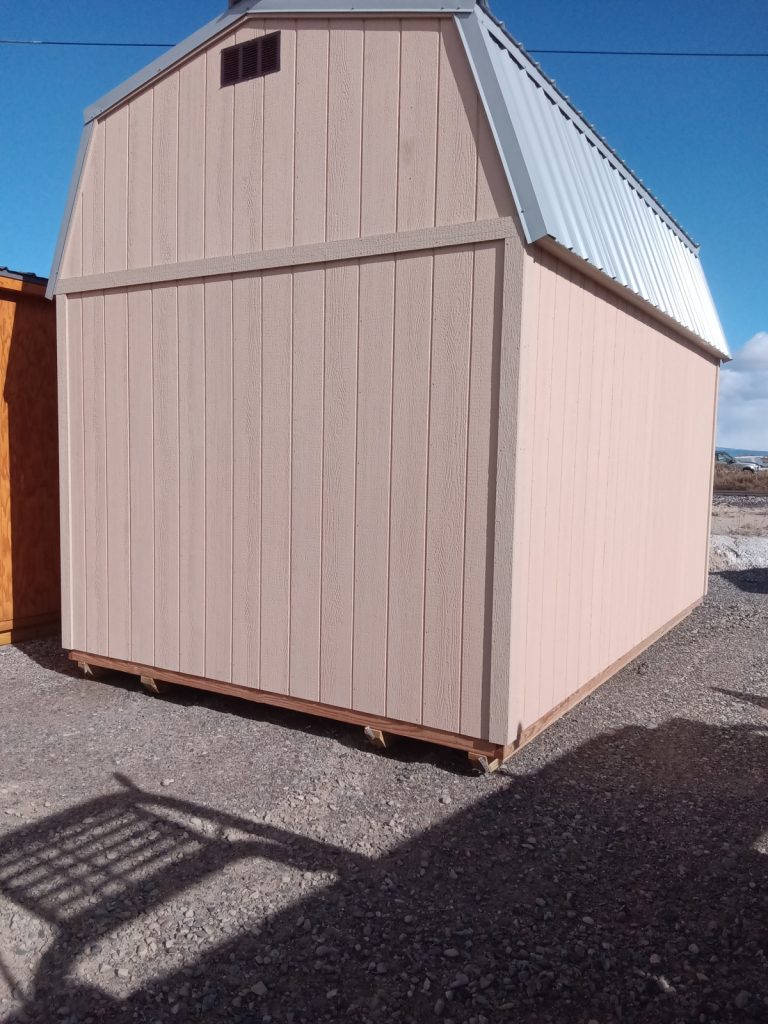 A Lofted Dutch Barn - 2368 featuring a tan wooden facade and a silver metal roof sits on a gravel surface beneath a clear blue sky. Shadows of a fence elegantly stretch across the ground in front, enhancing its rustic charm.