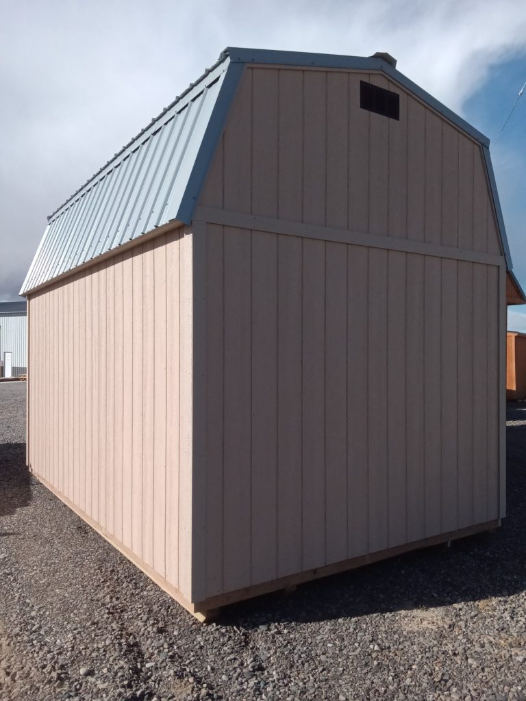 A Lofted Dutch Barn - 2368, small and light-colored, with a wooden structure and metal roof, stands on a gravel surface under a partly cloudy sky. It features the distinctive lofted design and includes a vent near the peak of the roof. Similar barns can be seen in the background, enhancing this picturesque rural setting.