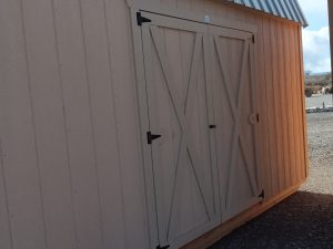The Lofted Dutch Barn - 2368, featuring a tan wooden exterior, metal roof, and X-braced double doors, exudes a charming rustic appeal. Positioned on a gravel base beneath an open sky, it stands alongside another partially visible structure to the right.