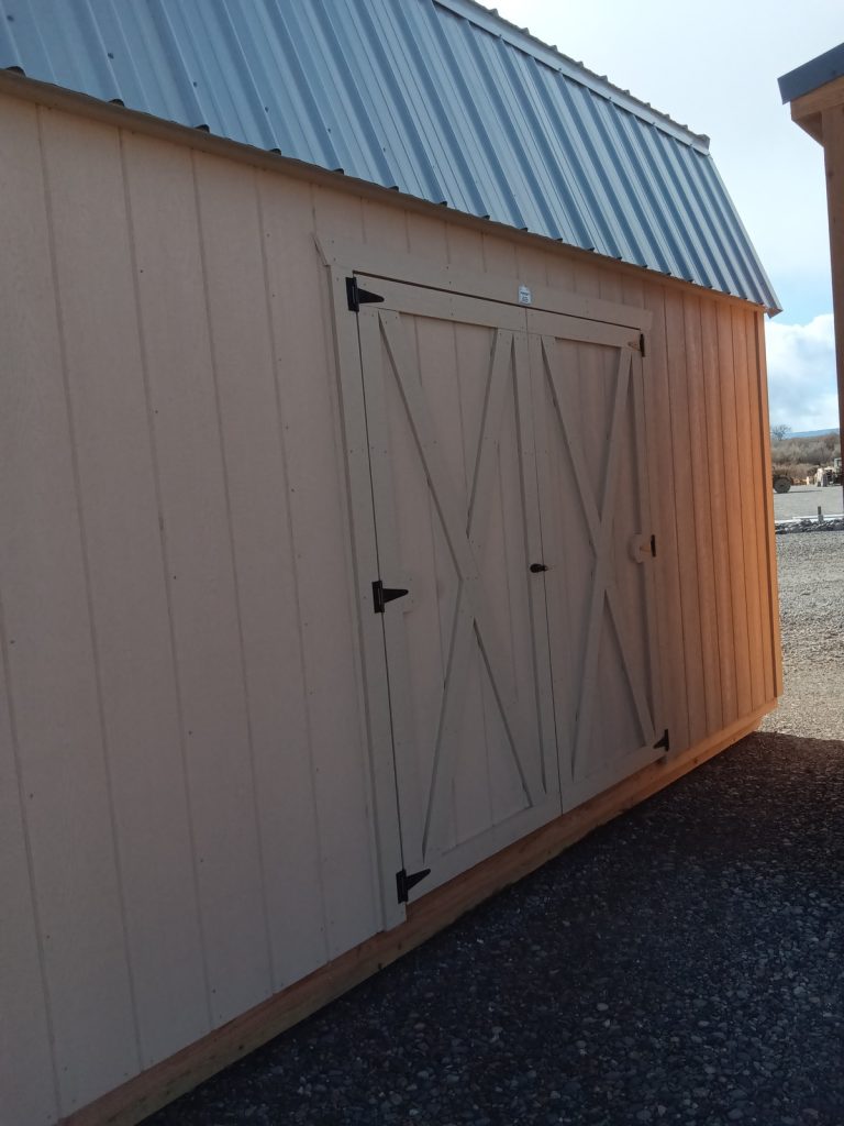 The Lofted Dutch Barn - 2368, featuring a tan wooden exterior, metal roof, and X-braced double doors, exudes a charming rustic appeal. Positioned on a gravel base beneath an open sky, it stands alongside another partially visible structure to the right.