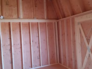 The Lofted Dutch Barn - 2368 showcases an unfinished interior with walls and a vaulted ceiling. The exposed plywood walls and floor, along with visible wooden studs, create a cohesive look. Soft natural light streams through a small upper window, enhancing its rustic charm.