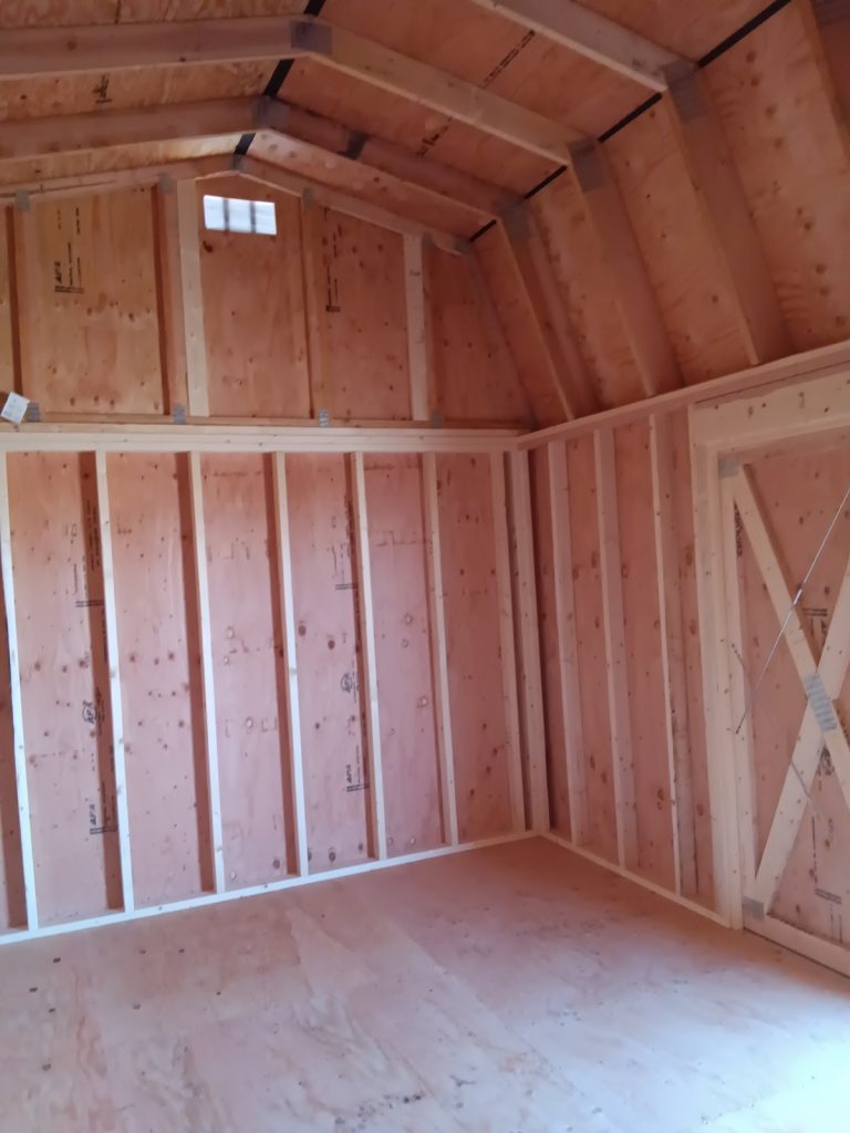 The Lofted Dutch Barn - 2368 showcases an unfinished interior with walls and a vaulted ceiling. The exposed plywood walls and floor, along with visible wooden studs, create a cohesive look. Soft natural light streams through a small upper window, enhancing its rustic charm.