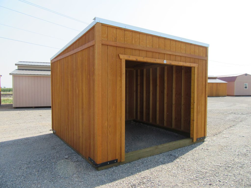 A wooden Buckaroo, open-front storage shed stands on a gravel lot. Its exterior consists of vertical wooden panels, and the interior is visible, showing a simple structure. Other similar sheds are seen in the background under a clear sky.