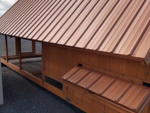 The Chicken House Large Roost - 2719 features a spacious wooden design with a slanted metal roof and rectangular extension, both topped with matching metal roofs, set on gravel under a cloudy sky.